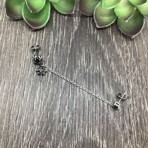 May include: A silver double-sided earring with black gemstones. The earring features a chain connecting two posts, each with a gemstone. One post has a floral design, and the other has a square gemstone.