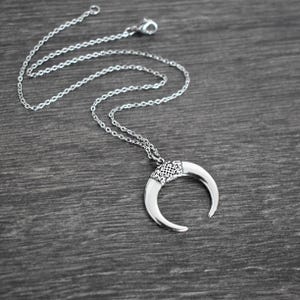May include: Silver-toned necklace featuring a crescent moon pendant. The pendant has a detailed, patterned top section and curved, horn-like ends. The chain is delicate and the necklace is displayed on a dark gray wooden surface.