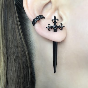 Pu&ograve; includere: Un orecchino nero con un design a forma di teschio e un piercing per orecchie nero con un design a forma di giglio e una lunga punta nera.