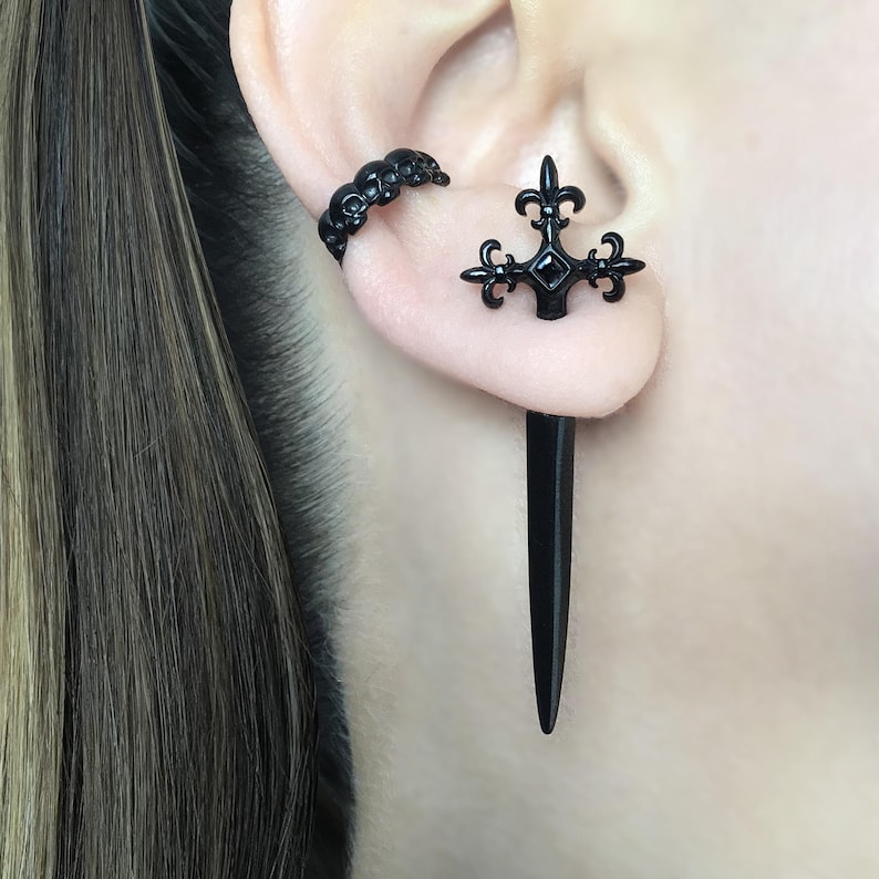 Pu&ograve; includere: Un orecchino nero con un design a forma di teschio e un piercing per orecchie nero con un design a forma di giglio e una lunga punta nera.