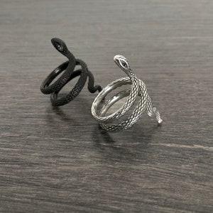 Puede incluir: Dos anillos con forma de serpiente, uno plateado y otro negro, sobre una superficie de madera oscura.