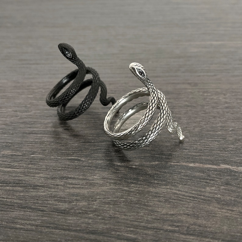 Puede incluir: Dos anillos con forma de serpiente, uno plateado y otro negro, sobre una superficie de madera oscura.