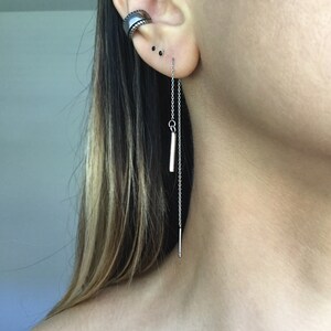 May include: A silver chain earring with a small silver bar pendant. The earring is hanging from the earlobe and the chain is long enough to reach the neck.