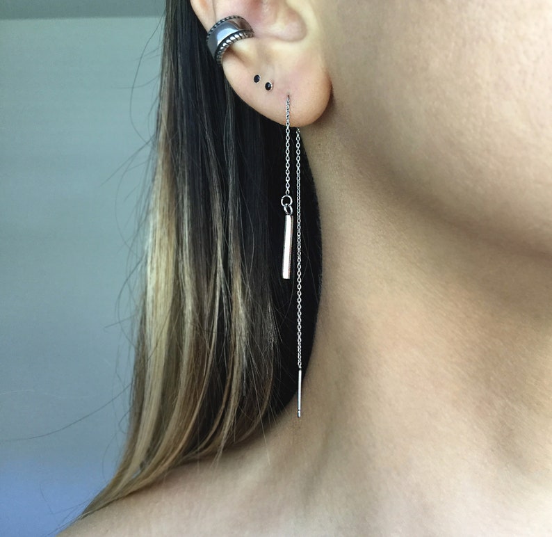 May include: A silver chain earring with a small silver bar pendant. The earring is hanging from the earlobe and the chain is long enough to reach the neck.
