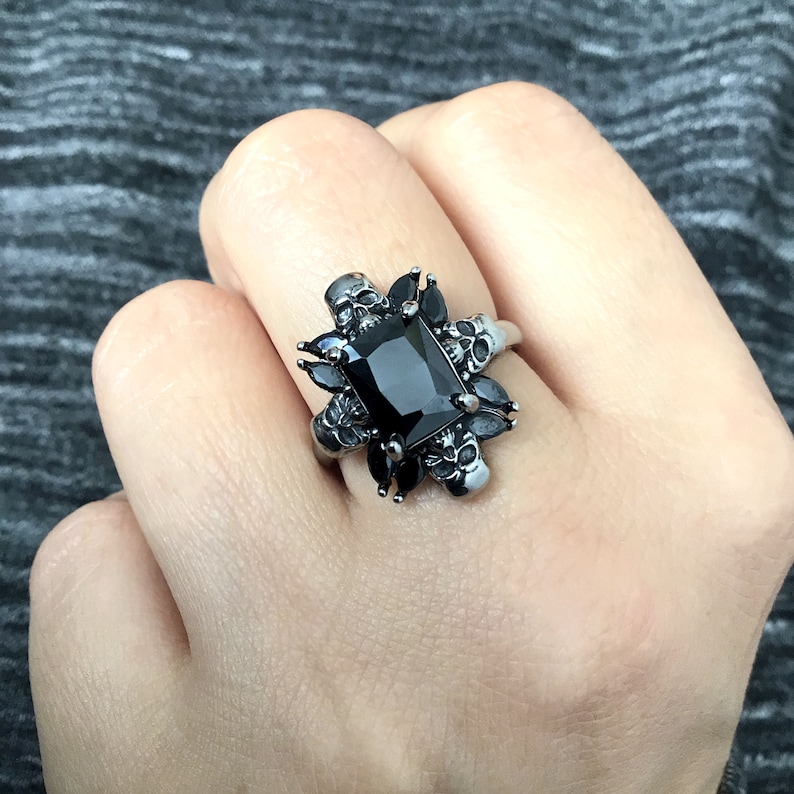 May include: A black onyx ring with a square-shaped stone surrounded by silver skull details.