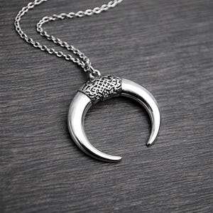 May include: Silver-toned crescent moon pendant necklace. The pendant has a textured band and a curved, polished horn shape. The chain is a delicate link style. The necklace is displayed on a dark wood-grain surface.