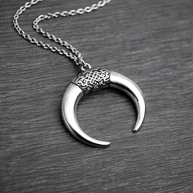 May include: Silver-toned crescent moon pendant necklace. The pendant has a textured band and a curved, polished horn shape. The chain is a delicate link style. The necklace is displayed on a dark wood-grain surface.