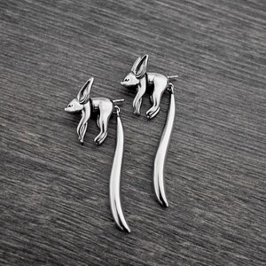 May include: A pair of silver dangle earrings featuring a stylized fennec fox design with long, curved tails.