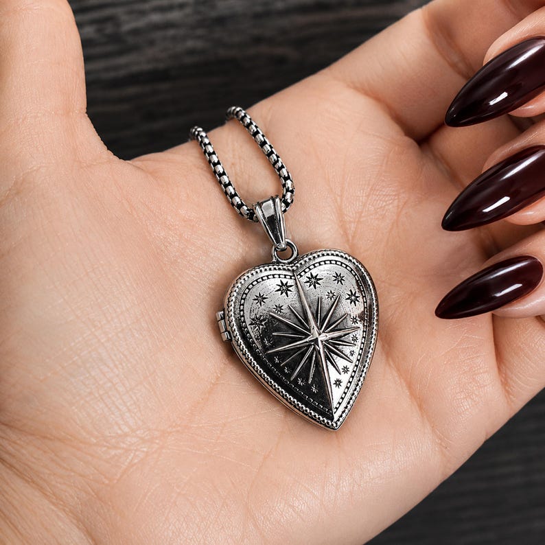 May include: A silver-toned heart-shaped locket pendant suspended from a textured box chain. The locket features an engraved North Star design surrounded by smaller star motifs and a beaded border, finished with an oxidised, darkened metal texture.