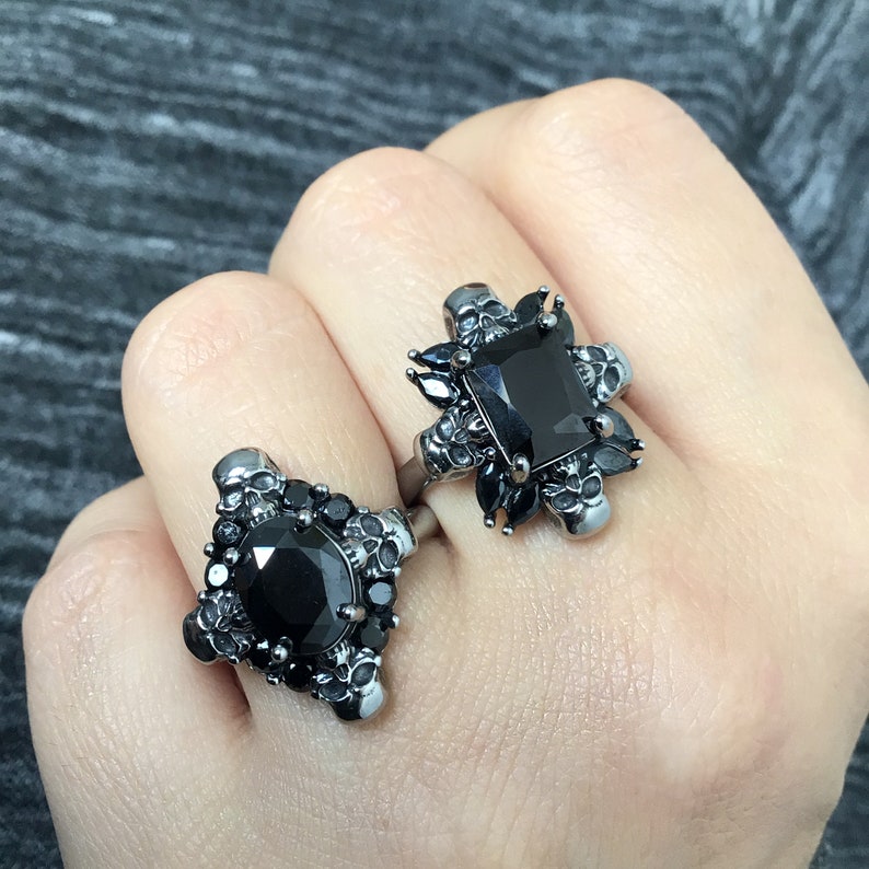 May include: Two black rings with skull and black gemstone accents. The rings are on a hand.