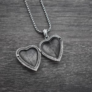 May include: A silver-toned heart-shaped locket pendant with an open design, featuring a textured interior and a beaded border. The locket is attached to a matching silver-toned box chain necklace, showcasing a metallic finish and intricate detailing.