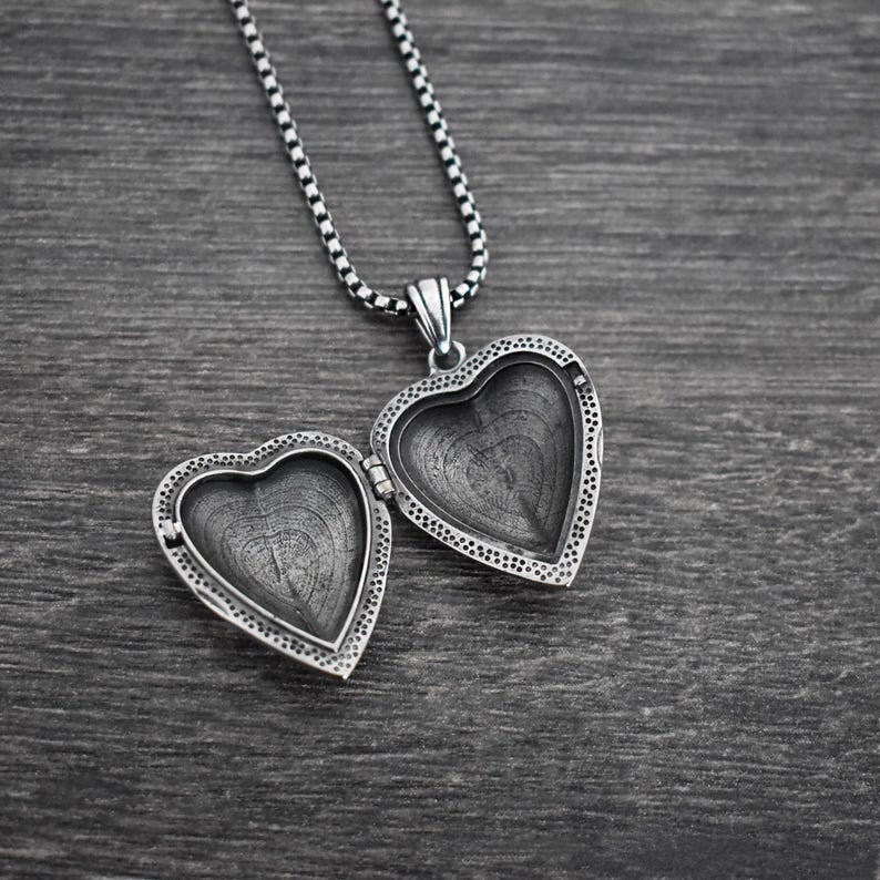May include: A silver-toned heart-shaped locket pendant with an open design, featuring a textured interior and a beaded border. The locket is attached to a matching silver-toned box chain necklace, showcasing a metallic finish and intricate detailing.