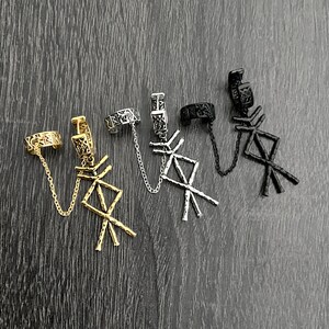 May include: Four ear cuff earrings in gold, silver, and black. Each cuff features a chain that connects to a dangling rune symbol. The earrings are displayed on a dark wood-grain surface.
