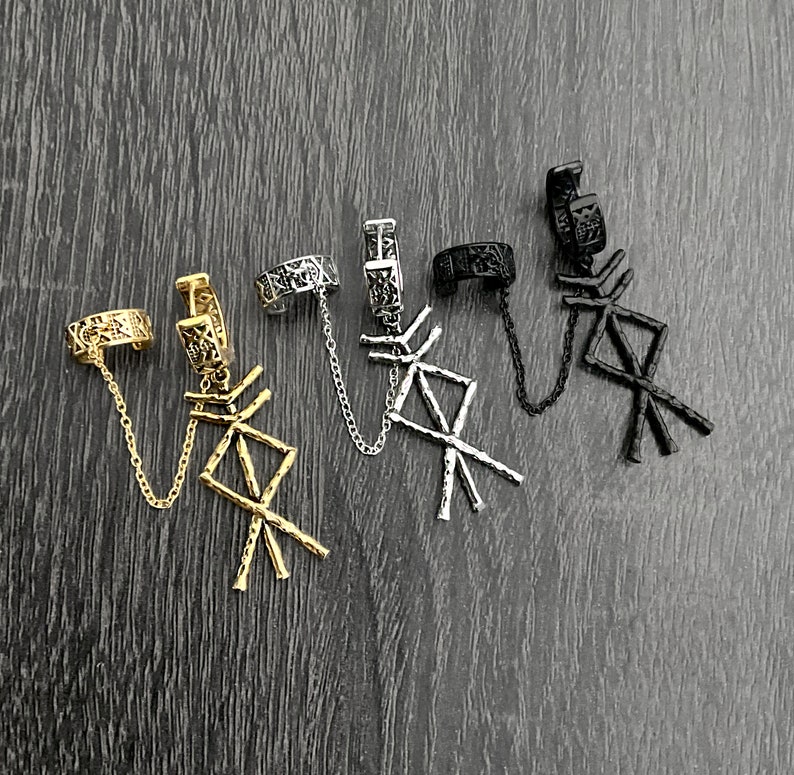 May include: Four ear cuff earrings in gold, silver, and black. Each cuff features a chain that connects to a dangling rune symbol. The earrings are displayed on a dark wood-grain surface.
