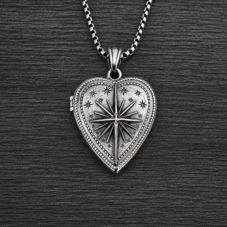May include: A silver-toned heart-shaped locket pendant suspended from a matching box chain. The locket features an embossed North Star design at the centre, surrounded by small engraved stars and a decorative beaded border with an oxidised finish.