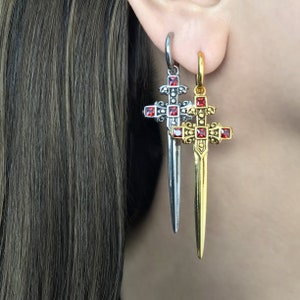 May include: A pair of earrings featuring a silver and gold sword cross pendant with red gemstone accents.