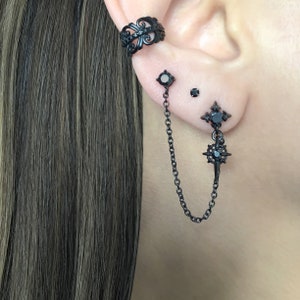 May include: Black ear cuff and earrings set. The ear cuff features an ornate design. The earrings include a stud, a small round stud, and a double-piercing earring with a chain and starburst charm. The jewelry is black.