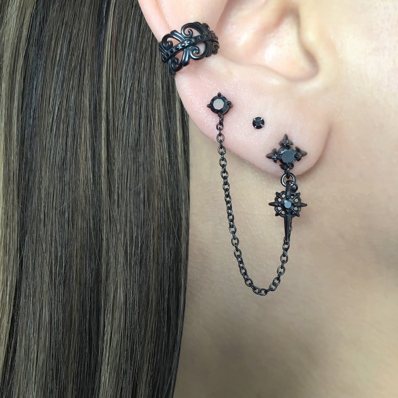 May include: Black ear cuff and earrings set. The ear cuff features an ornate design. The earrings include a stud, a small round stud, and a double-piercing earring with a chain and starburst charm. The jewelry is black.