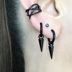 May include: A set of black jewelry consisting of a thorn-patterned ear cuff, two hoop earrings with dangling black spikes, and a small black stud earring. The pieces are made of a smooth, dark material and feature a consistent matte black finish.