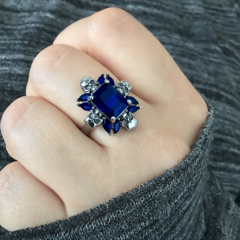 May include: A silver ring featuring a large blue gemstone surrounded by smaller blue gemstones and silver skull accents.