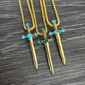 May include: Three gold necklaces with sword pendants. Each sword is adorned with blue and green gemstones.