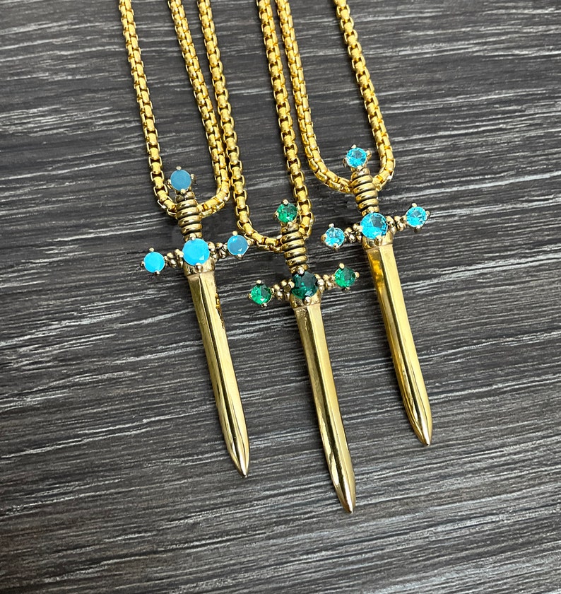 May include: Three gold necklaces with sword pendants. Each sword is adorned with blue and green gemstones.