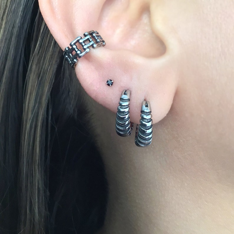 May include: A close-up of a person's ear with three silver earrings. The top earring is a chain link design, the middle earring is a small black stud, and the bottom two earrings are silver hoops with a textured design.