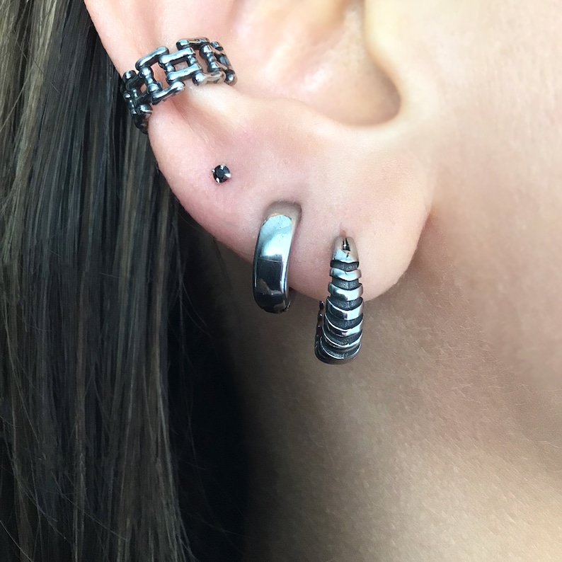 May include: A person's ear with three silver earrings. The top earring is a chain link design, the middle earring is a small black stud, and the bottom earring is a silver hoop with a textured design.