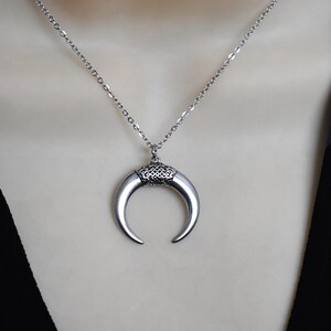 May include: A silver-toned crescent moon pendant necklace. The pendant has a curved horn shape with a decorative top. The necklace chain is delicate and also silver-toned. The necklace is displayed on a neutral background.