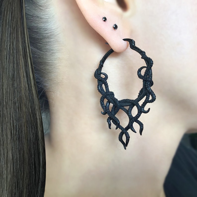 May include: Black hoop earrings with a detailed design resembling a cluster of branches or vines. The earrings are made of a dark, matte material and have a slightly textured finish.