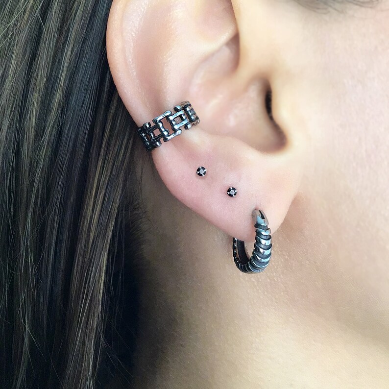 May include: A person's ear with three black metal earrings. The top earring is a chain link design, the middle two are small studs with black stones, and the bottom earring is a thick hoop with a textured design.