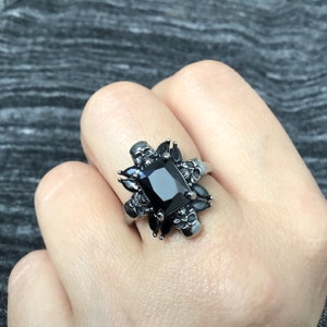May include: A black onyx ring with a square-shaped stone surrounded by silver skulls. The ring is set in a silver band.