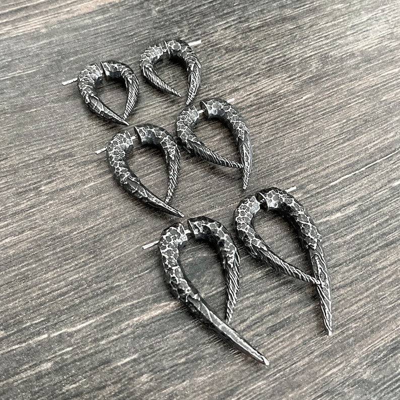 Peut inclure: Cinq paires de boucles d'oreilles en m&eacute;tal noir avec une texture martel&eacute;e et une forme pointue et incurv&eacute;e. Les boucles d'oreilles sont dispos&eacute;es en quinconce sur une surface en bois.