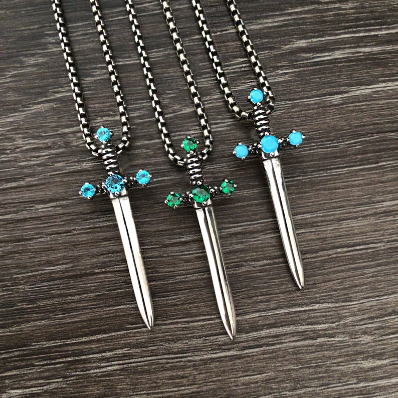 May include: Three silver sword pendants with chains. The swords have blue and green gemstones.