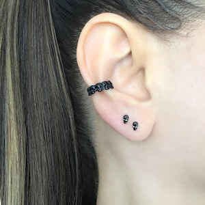 May include: Black ear cuff and skull stud earrings. The ear cuff features a row of small skulls. The stud earrings are tiny black skulls. The jewelry is worn on the ear.