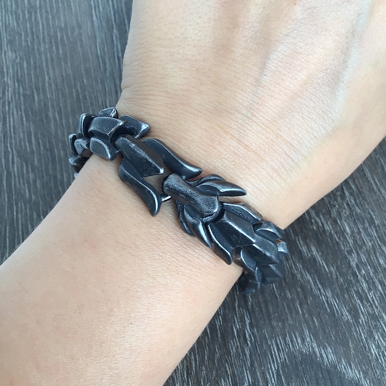 Puede incluir: Un brazalete de metal negro con un dise&ntilde;o &uacute;nico e intrincado. El brazalete est&aacute; hecho de piezas enlazadas e interconectadas que se asemejan a una cadena.