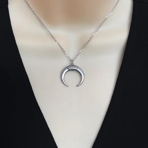 May include: A silver-toned necklace featuring a crescent moon pendant. The pendant has a textured top and curved horns. The necklace is displayed on a mannequin with a black top.