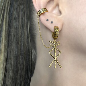 May include: Gold-toned ear cuff and earring set. The ear cuff wraps around the upper ear and connects to a hoop earring with a chain. The earring features a dangling, textured, geometric pendant. The jewelry has an engraved design.