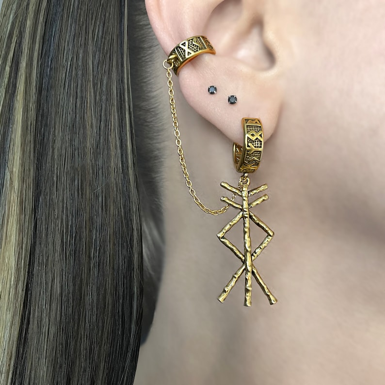 May include: Gold-toned ear cuff and earring set. The ear cuff wraps around the upper ear and connects to a hoop earring with a chain. The earring features a dangling, textured, geometric pendant. The jewelry has an engraved design.