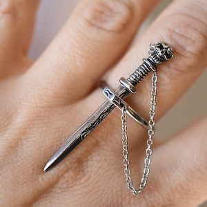 Könnte beinhalten: Ein silberner Schwertring mit einem Totenkopfdetail am Knauf und einer baumelnden Kette. Der Ring wird an einem Finger getragen.