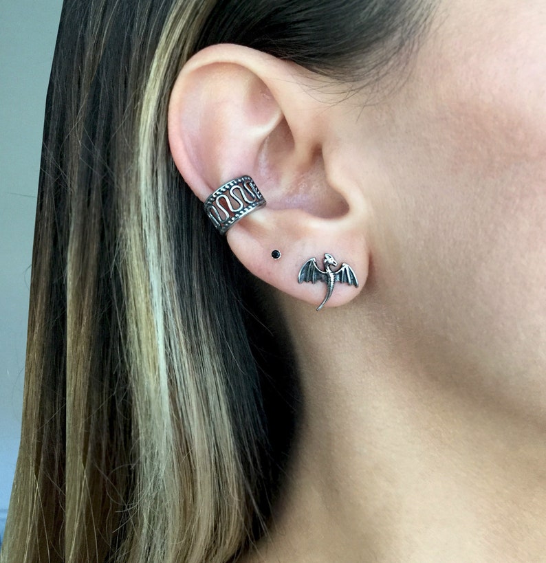May include: A silver ear cuff with a decorative design, a small black stud earring, and a silver dragon stud earring.