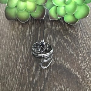May include: A silver snake ring with a coiled body and a detailed head. The ring is on a wooden surface.