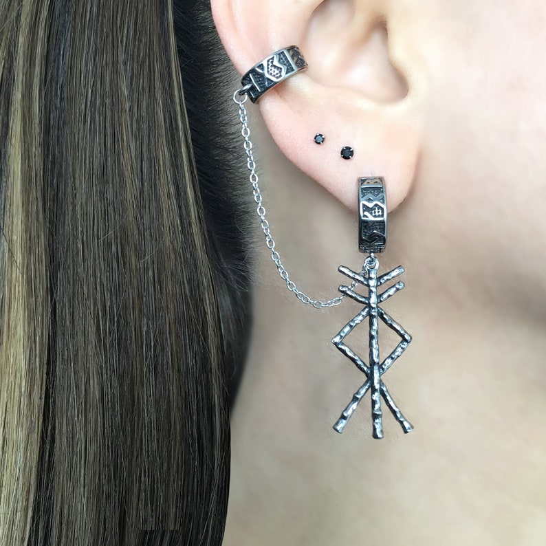 May include: Silver ear cuff and earring set. The ear cuff wraps around the upper ear and connects to a dangling earring with a chain. The earring features a geometric design. The jewelry is worn on a person's ear.