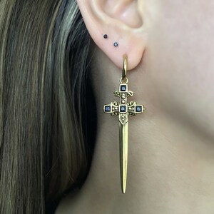 May include: Gold sword cross earring with black gemstone accents. The earring is hanging from a person's ear.