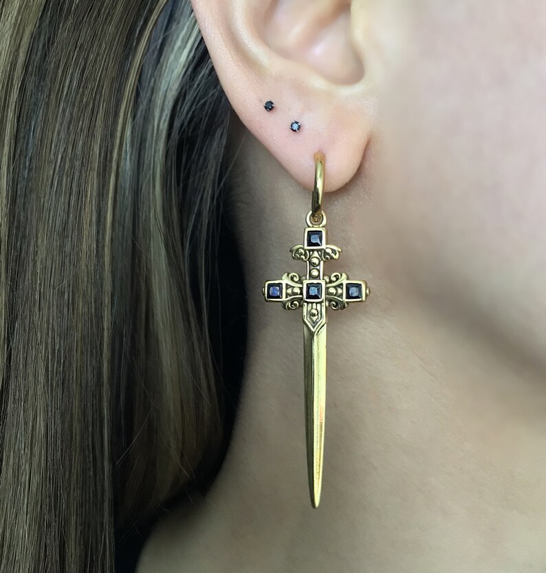 May include: Gold sword cross earring with black gemstone accents. The earring is hanging from a person's ear.