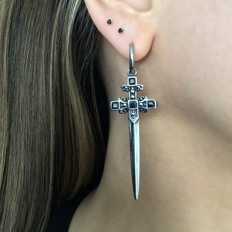 May include: A silver sword-shaped earring with a black cross pendant and black gemstones. The earring is hanging from a person's ear.