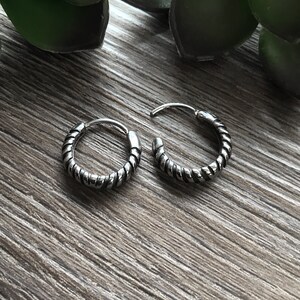 May include: Two silver hoop earrings with a twisted rope design. The earrings are on a wooden surface.