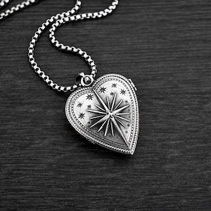 May include: A silver-toned heart-shaped locket pendant featuring an embossed North Star design surrounded by small stars and a beaded border. The locket is attached to a silver-toned box chain necklace with a polished finish and dark oxidised details.