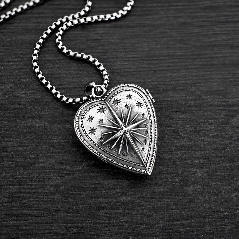 May include: A silver-toned heart-shaped locket pendant featuring an embossed North Star design surrounded by small stars and a beaded border. The locket is attached to a silver-toned box chain necklace with a polished finish and dark oxidised details.