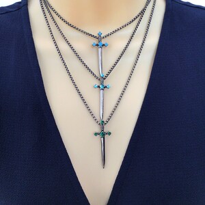 May include: Layered silver chain necklace with three sword pendants. The swords have turquoise and green gemstones.